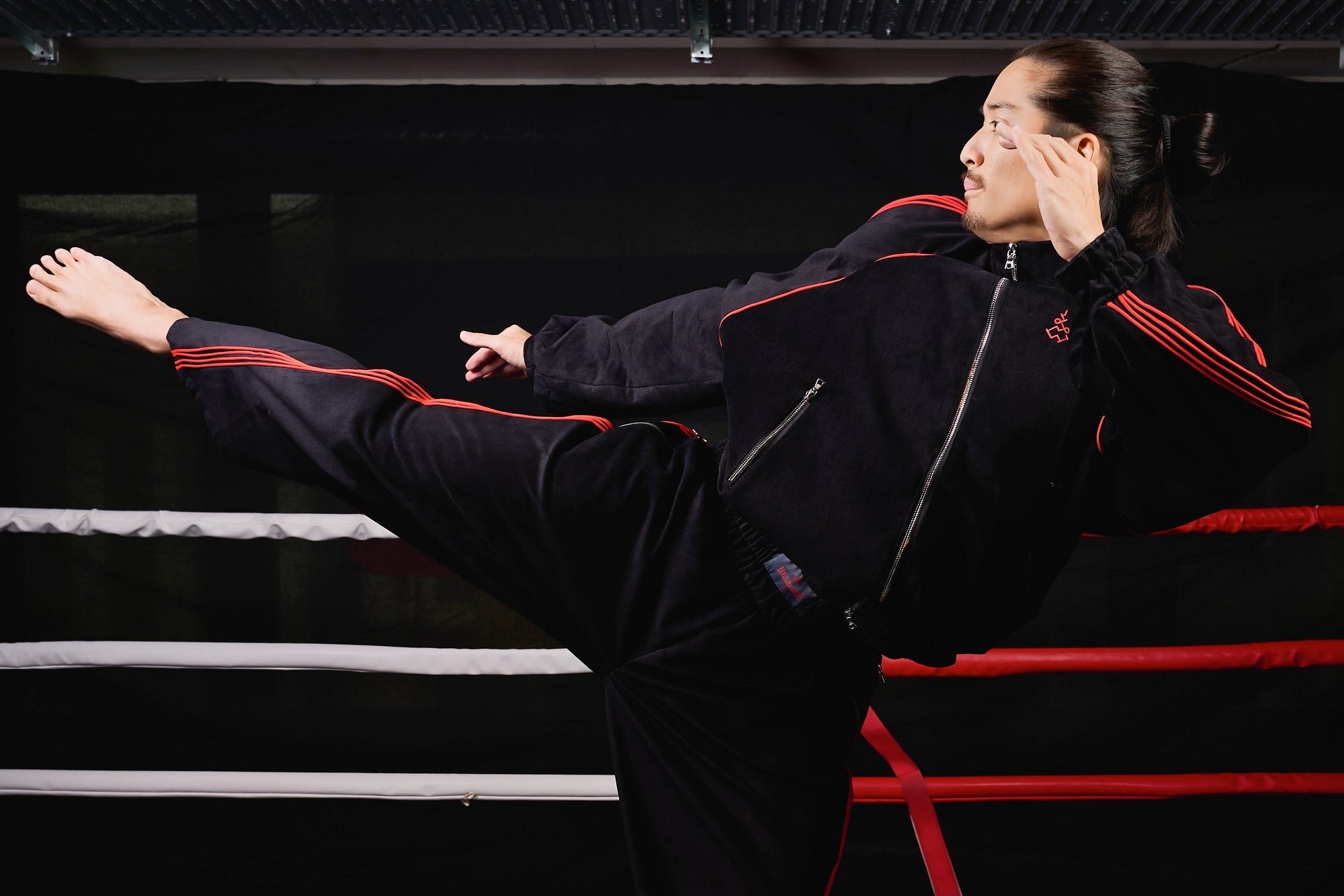 A man in a black athletic tracksuit with red stripes performs a high kick inside a boxing ring at a martial arts gym. Muay Thai Streetwear Sak Yant