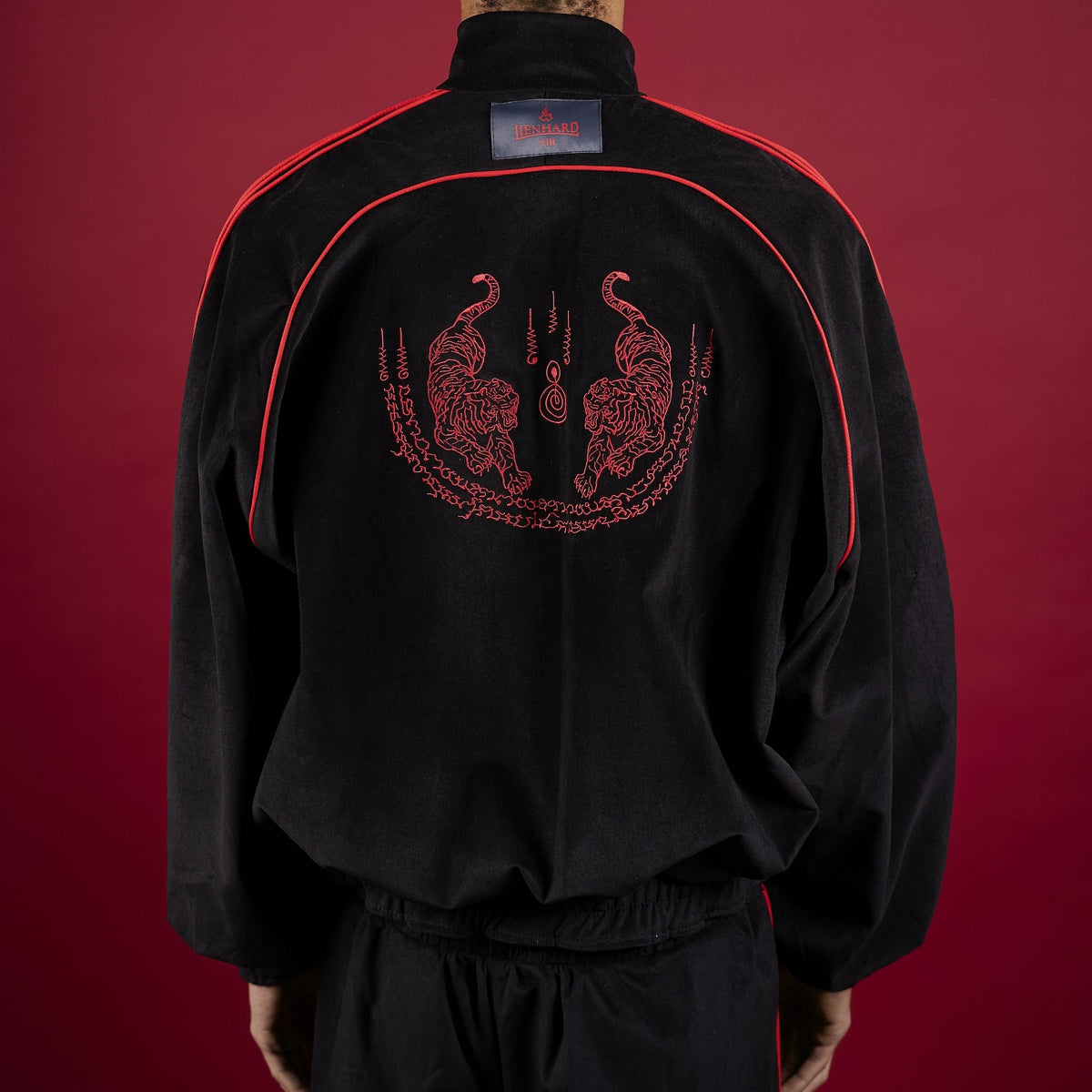 Person wearing a black tracksuit with red accents on a red background Lienhard VIII Muay Thai Streetwear Jacket Tracksuit Sak Yant Jacket