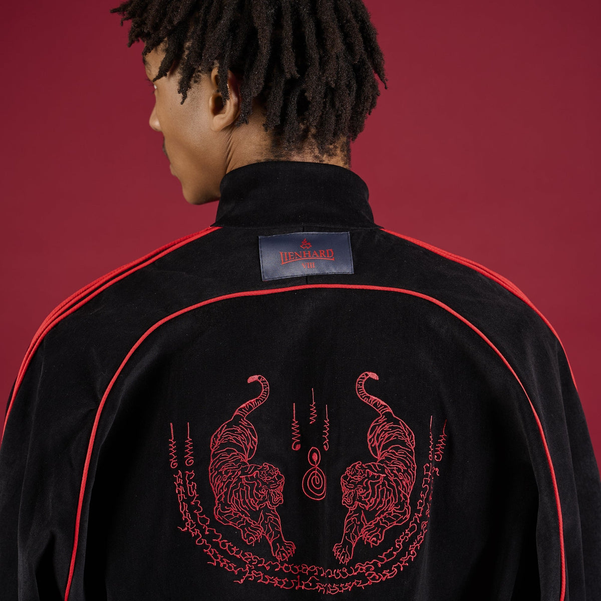 Person wearing a black tracksuit with red accents on a red background Lienhard VIII Muay Thai Streetwear Jacket Tracksuit Sak Yant Jacket