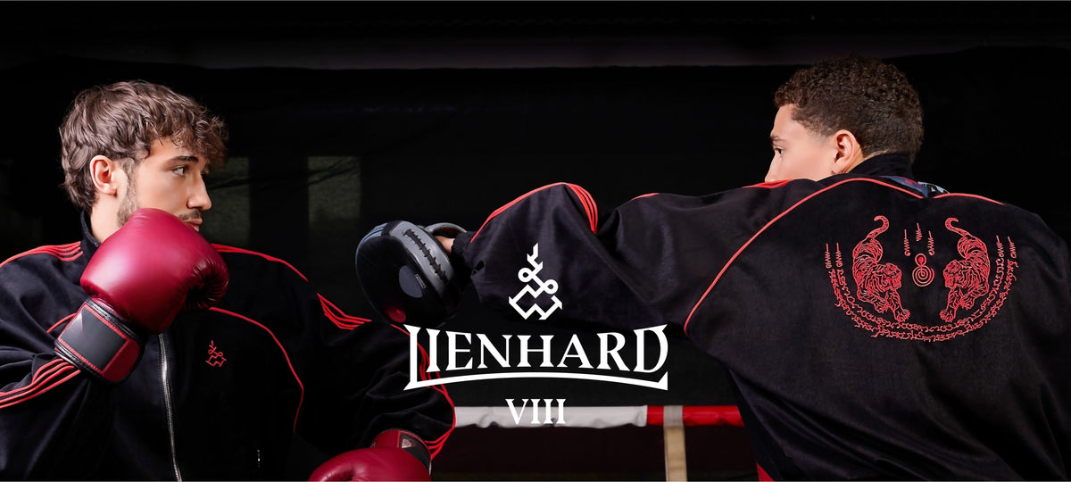 Two men in black martial arts uniforms with red trim and red boxing gloves sparring in a dark indoor setting. The back of the uniform on the right features a red dragon emblem, and the center of the image has a prominent 'LIENHARD VIII' logo overlay. Muay Thai Streetwear Sak Yant Tattoo Thai Boxing Tracksuit Muay Thai
